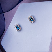 A pair of turquoise power embellished stud earrings placed on a white surface with a blue textured background.